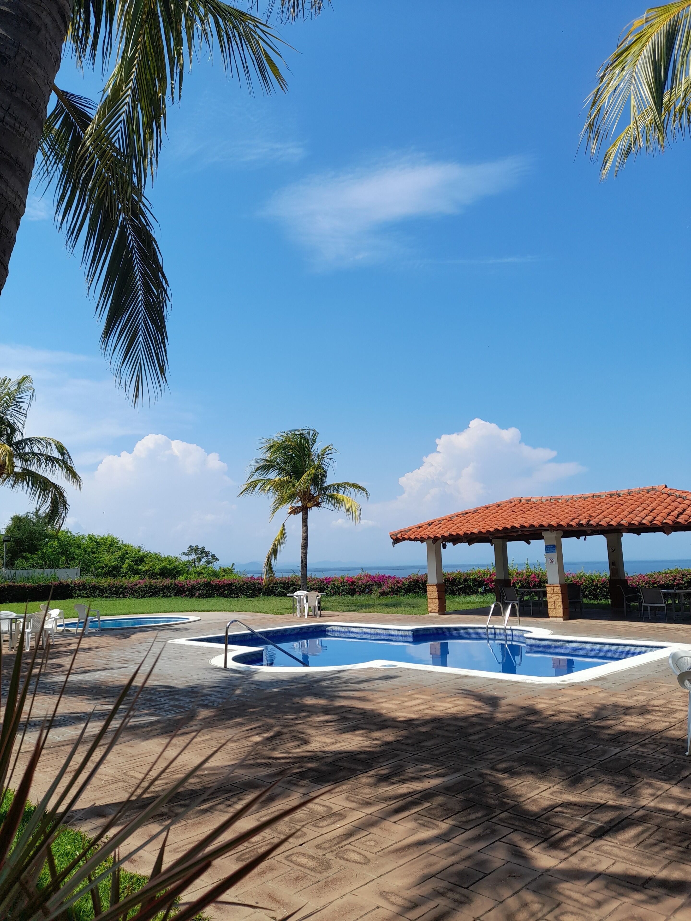 Uma área de piscina com um deck ladrilhado, um espaço coberto para sentar e vista para o oceano.