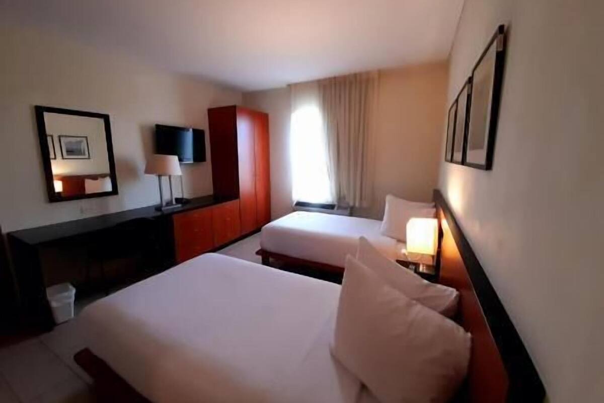 A hotel room with two beds, a desk, a TV, and a window with curtains.