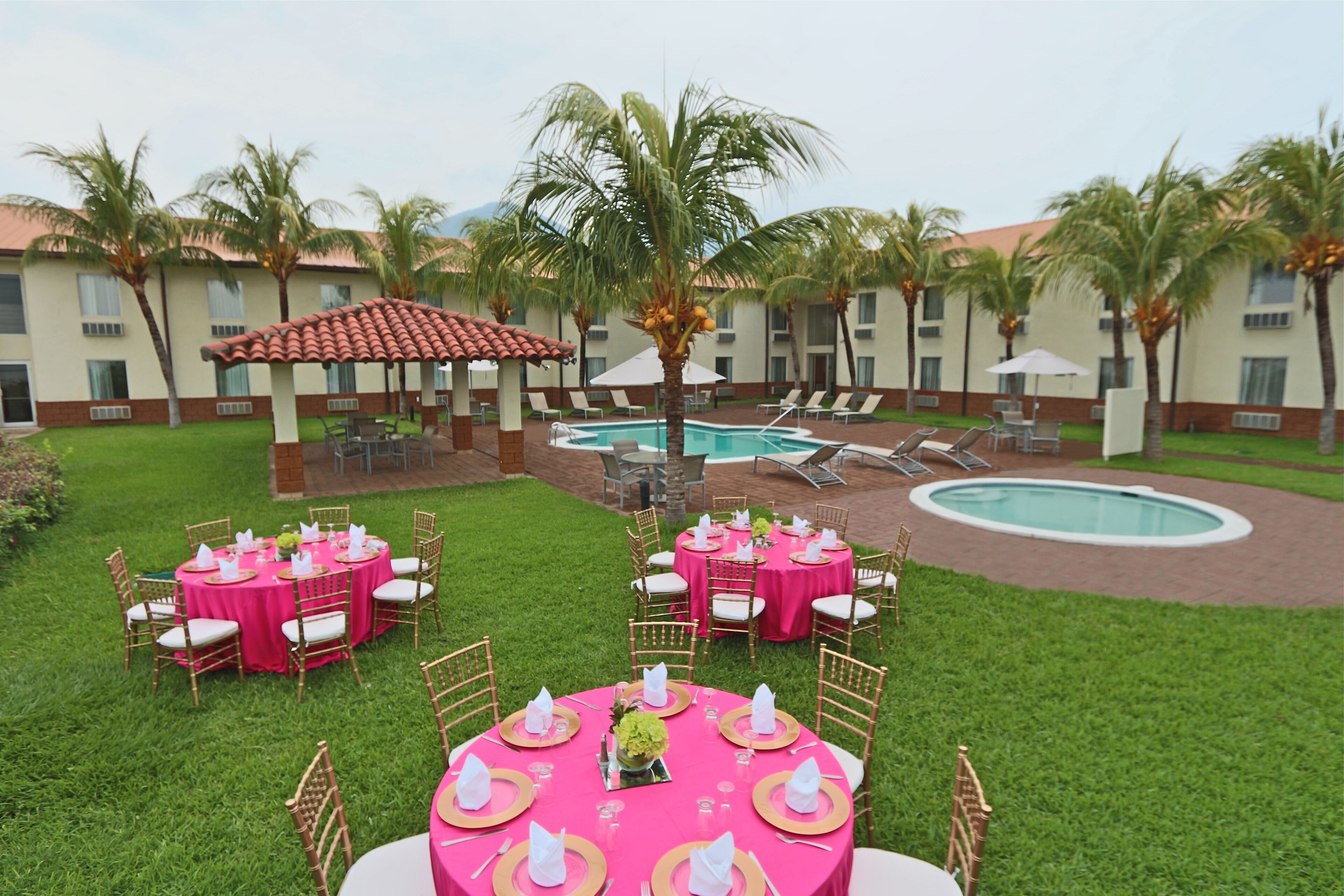Un espacio de comedor al aire libre con manteles rosas y sillas blancas, una piscina y palmeras.