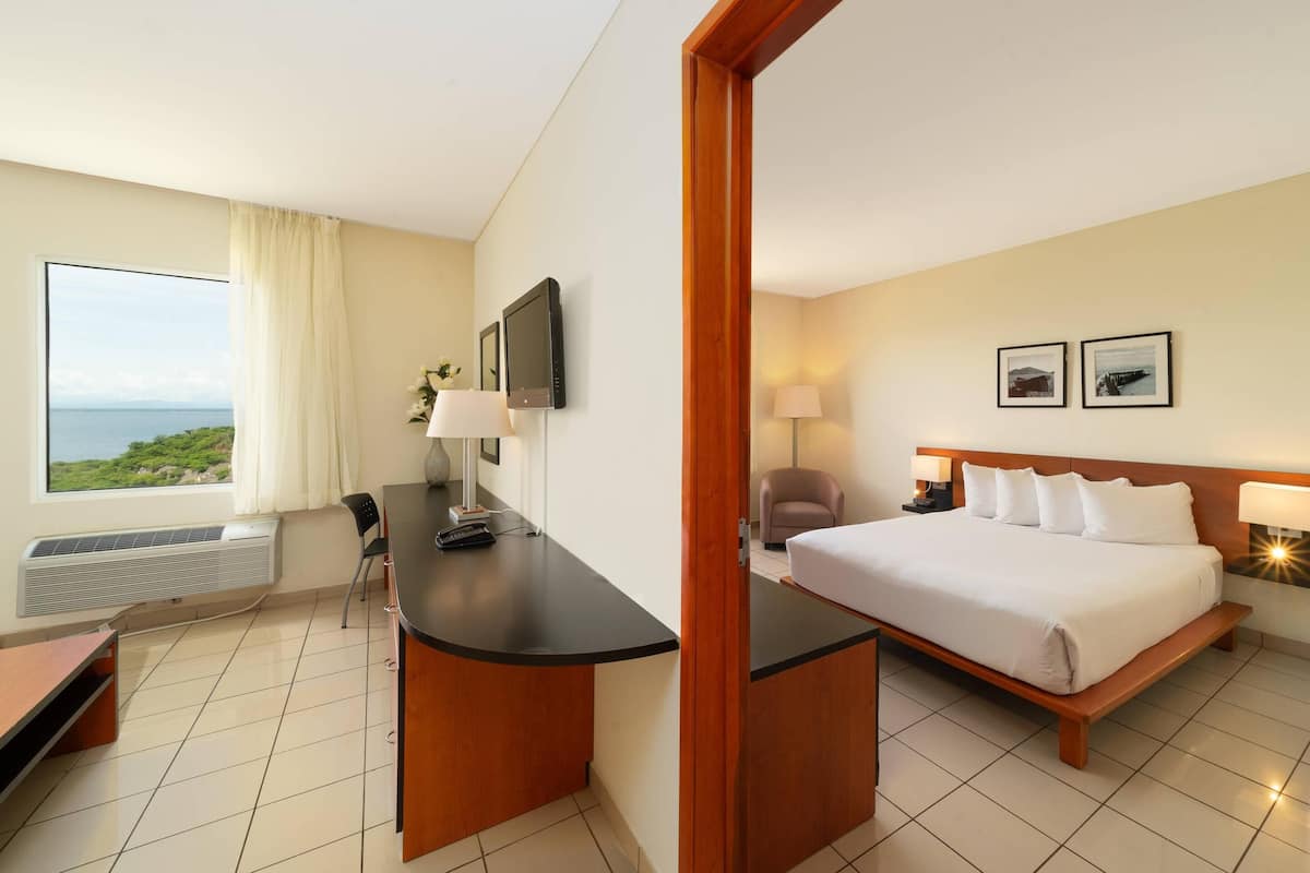 A hotel room with a bed, desk, chair, and a large window offering a view of the sea.
