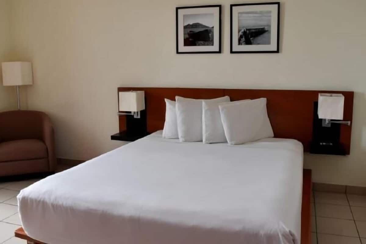 A hotel room with a bed, a chair, two framed pictures on the wall, and a nightstand with a lamp.