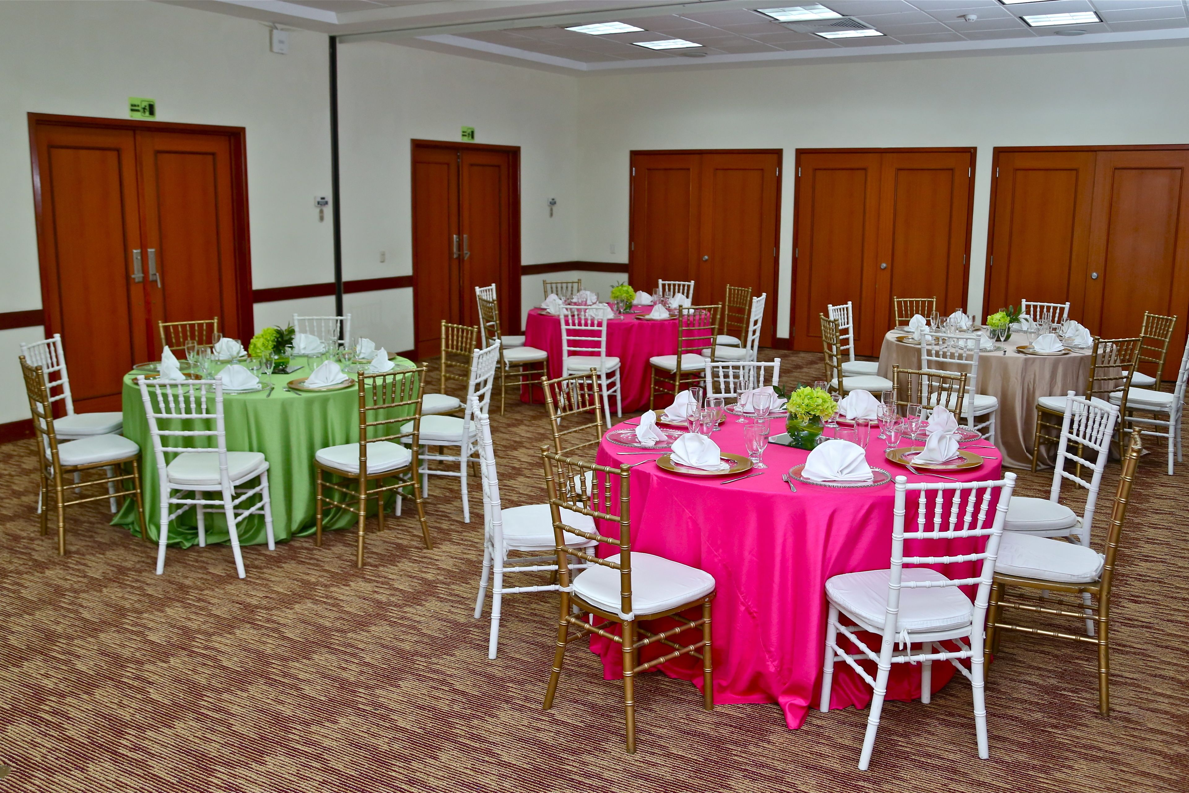 A banquet hall with multiple round tables, each with white chairs and covered with green or pink tablecloths.