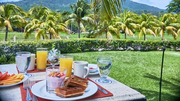 Una mesa de desayuno con diversas bebidas y alimentos, con palmeras y una montaña de fondo.