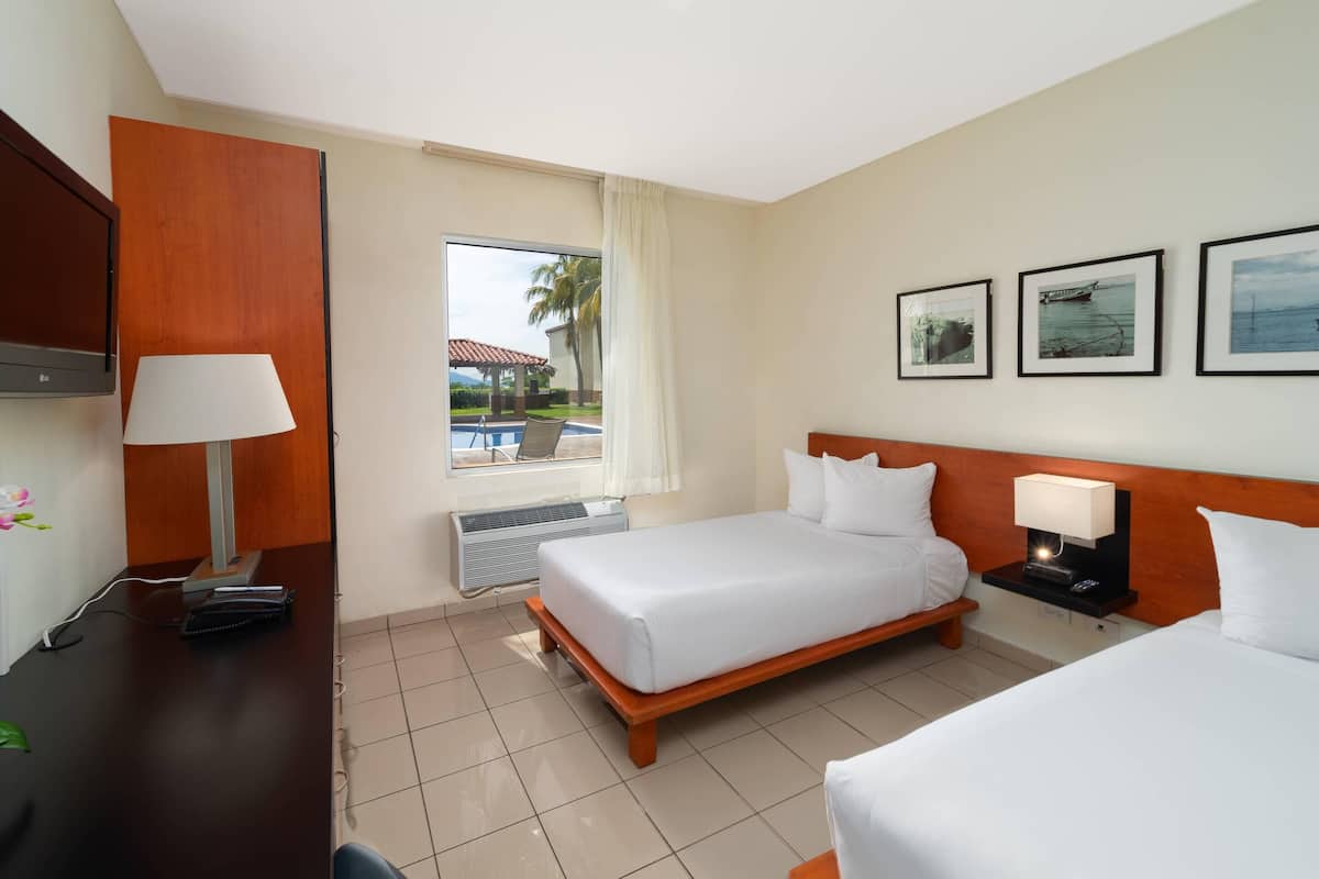 A hotel room with a bed, a desk, a chair, a TV, a window with a view of a pool, and framed pictures on the wall.