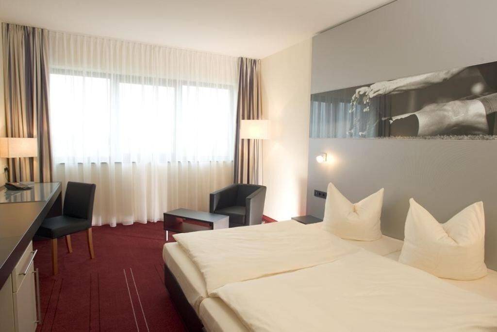 A hotel room with a large bed, two bedside tables, a desk, and a chair. There is a window with curtains and a wall-mounted artwork.