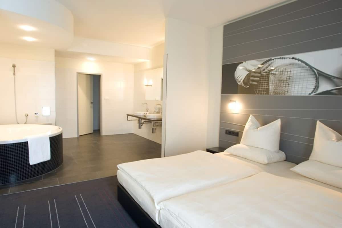 Een moderne hotelkamer met een groot bed, een aparte badkamer met badkuip, en een tennisracket als decoratie aan de muur.