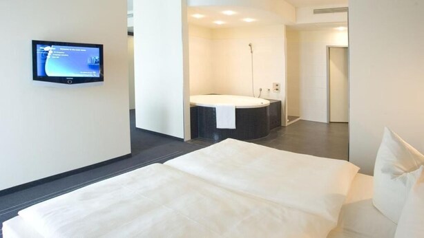 A modern hotel room with a large bed, a separate bathroom with a bathtub, and a flat-screen TV mounted on the wall.