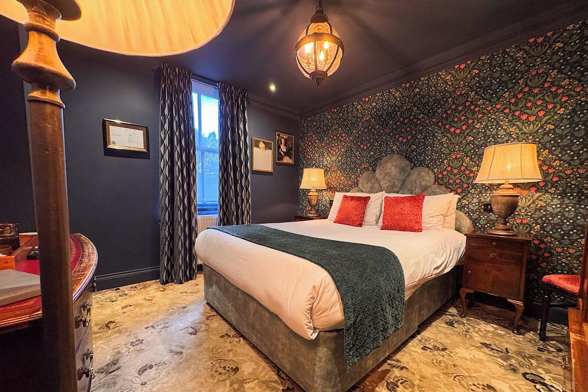 A bedroom with a large bed, a nightstand with a lamp, a dresser, and a window with curtains.