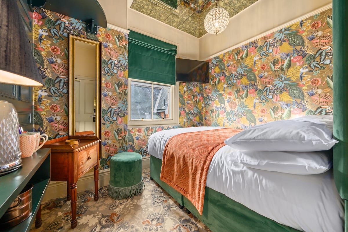 A bedroom with floral wallpaper, a green upholstered bench, a wooden dresser, and a bed with white and orange bedding.
