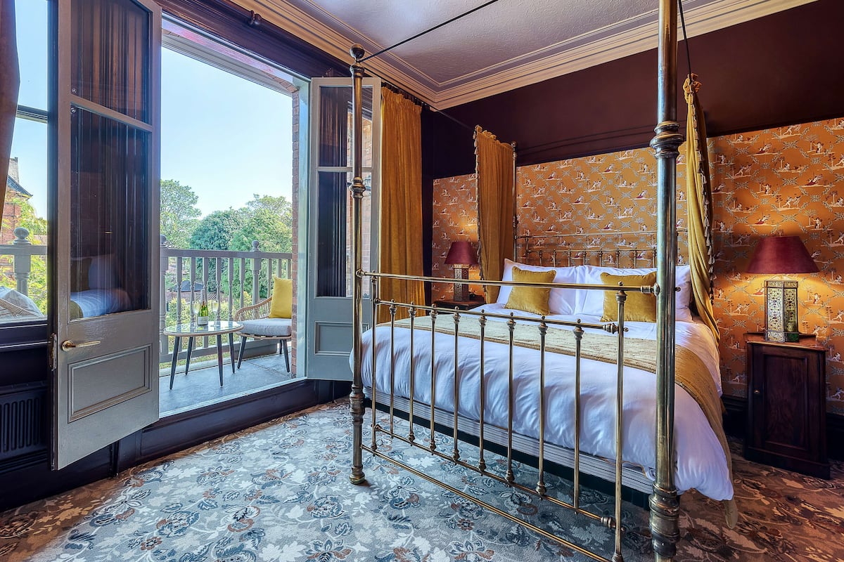 A four-poster bed with a canopy, a balcony with a view of greenery, and a patterned carpet.