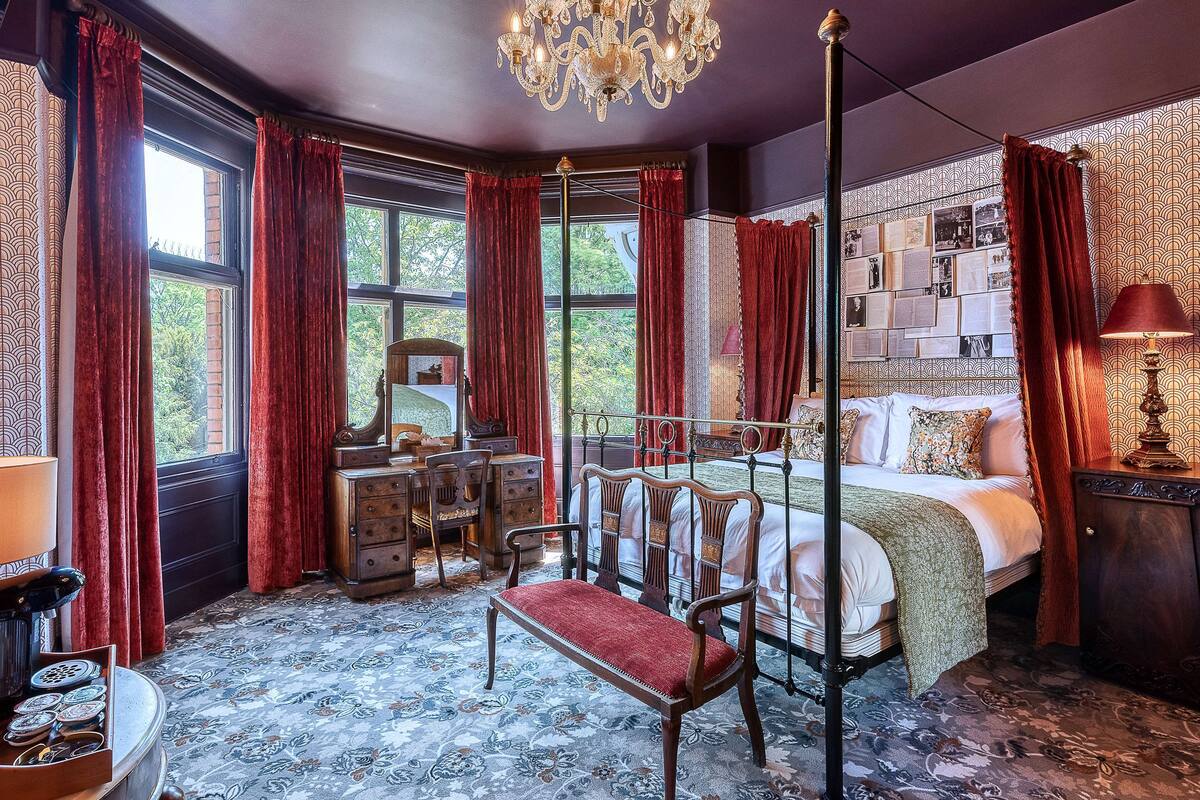 A bedroom with a four-poster bed, a chandelier, a desk with a chair, and a patterned carpet.