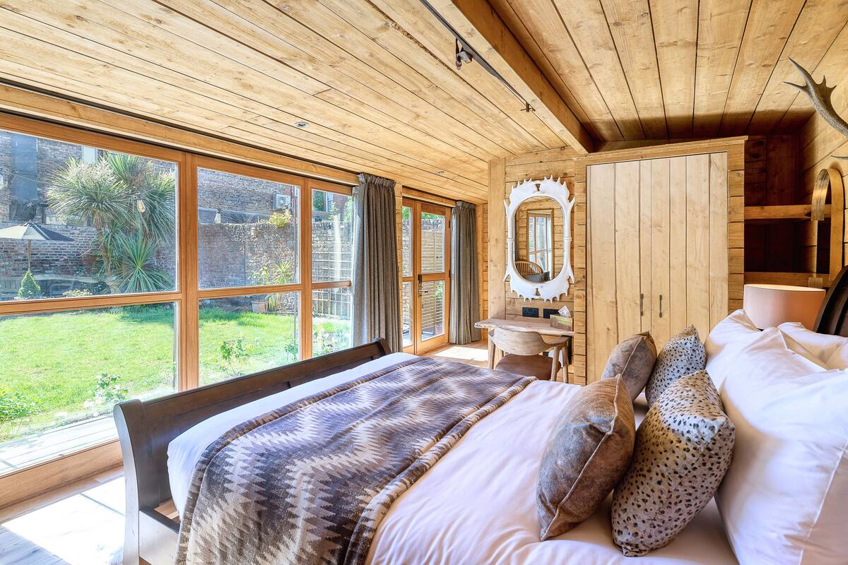 A bedroom with a large bed, a wooden wardrobe, a mirror, and a view of the garden.