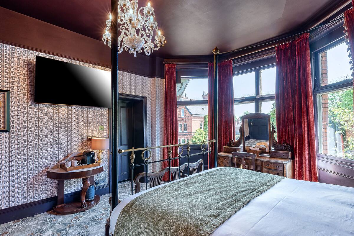 A hotel room with a large bed, a desk with a chair, a small round table, and a large window with curtains.