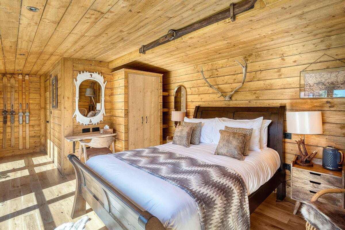 A wooden cabin bedroom with a bed, dresser, mirror, and wall-mounted lamp.