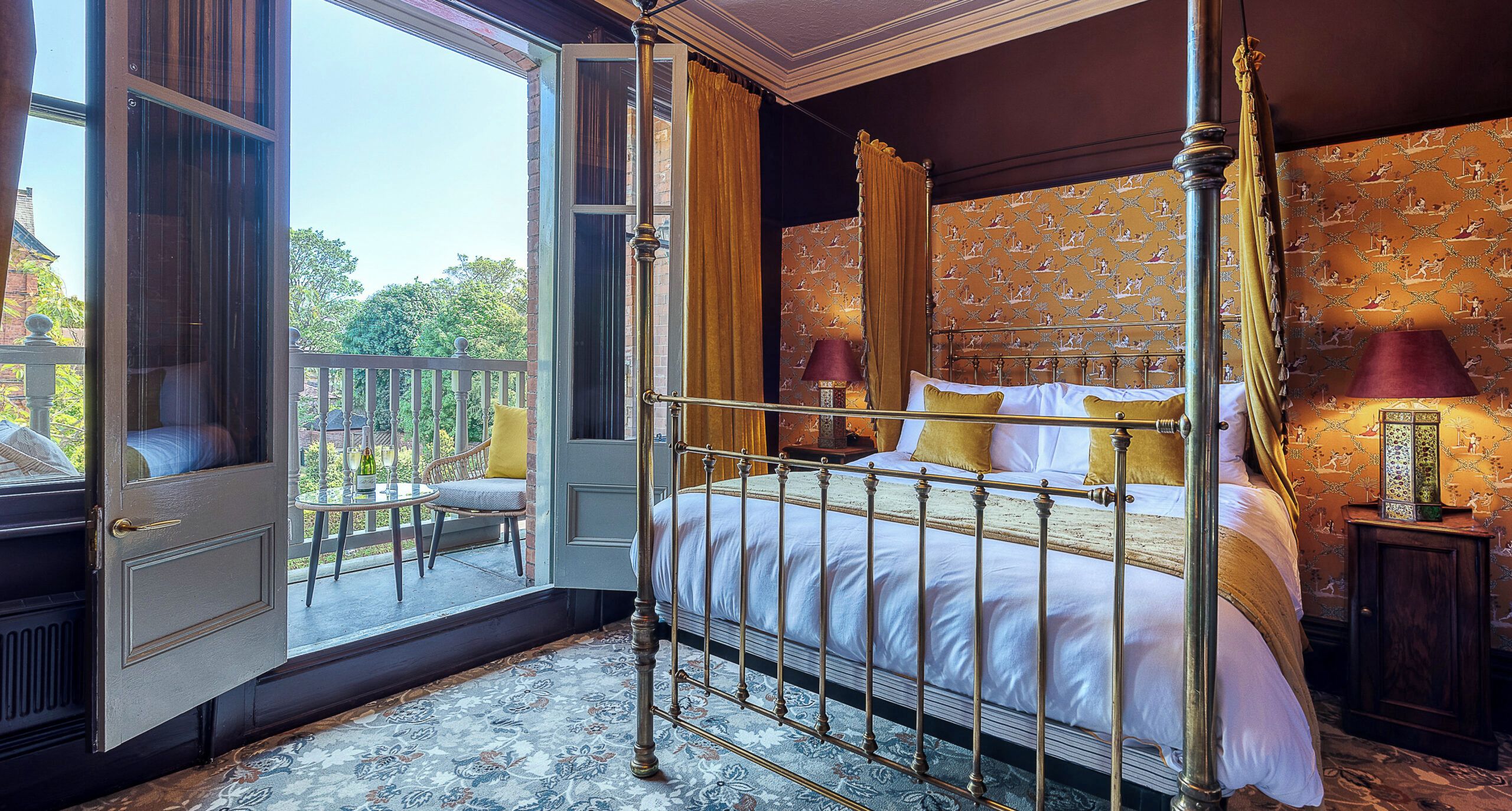 A four-poster bed with a canopy, a small table with a bottle and glasses, and a view of the outdoors through an open door.