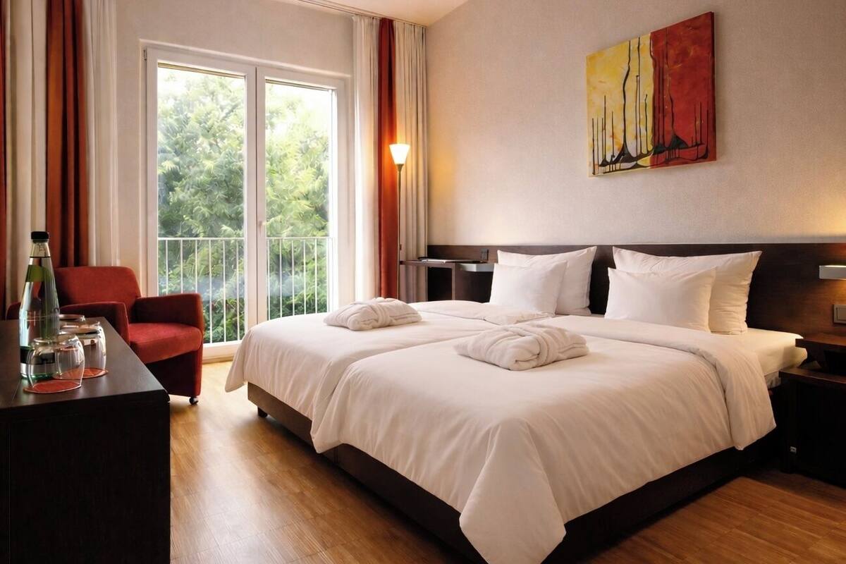 A hotel room with a large bed, a desk with a chair, a painting on the wall, and a balcony with curtains.
