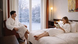 A hotel room with a large window, a bedside table, a reading lamp, and a painting on the wall.