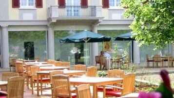 Un café al aire libre con mesas y sillas de madera, un área de estar techada, y un edificio con fachada de ladrillo rojo y detalles en blanco al fondo.