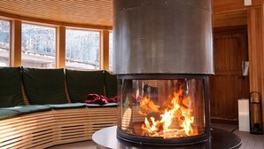 A modern interior with a central wood-burning stove, a curved seating area with cushions, and large windows providing a view of the outside.