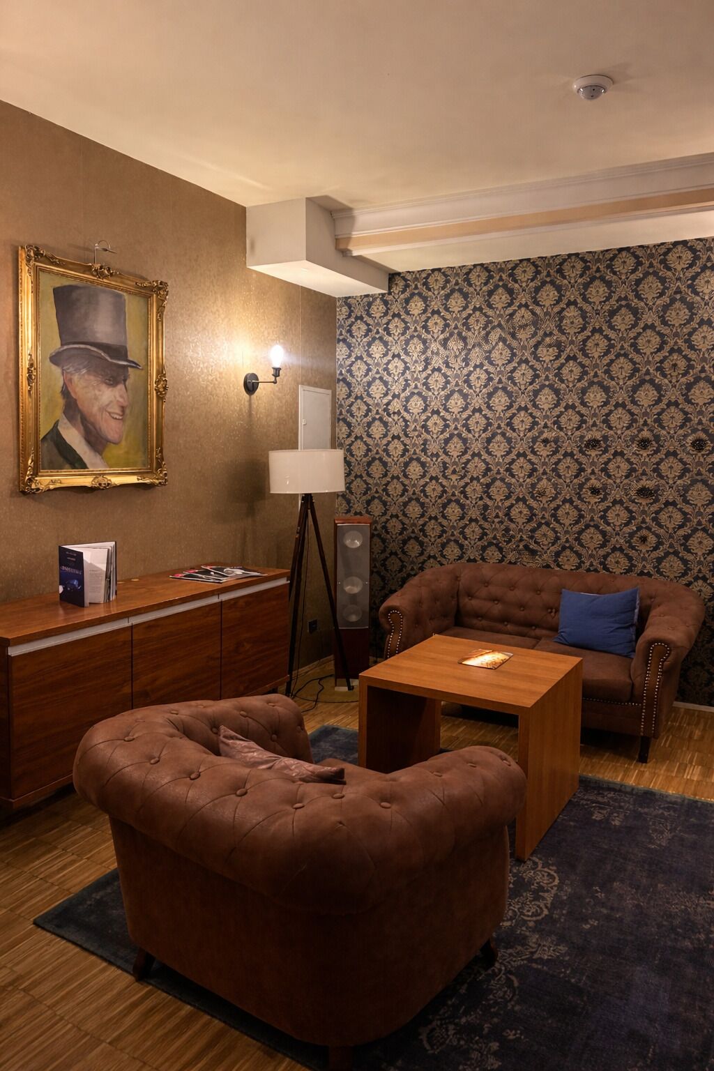 Una habitación acogedora con un sofá marrón, una mesa de centro de madera, una lámpara, un cuadro enmarcado en la pared y un papel tapiz estampado.