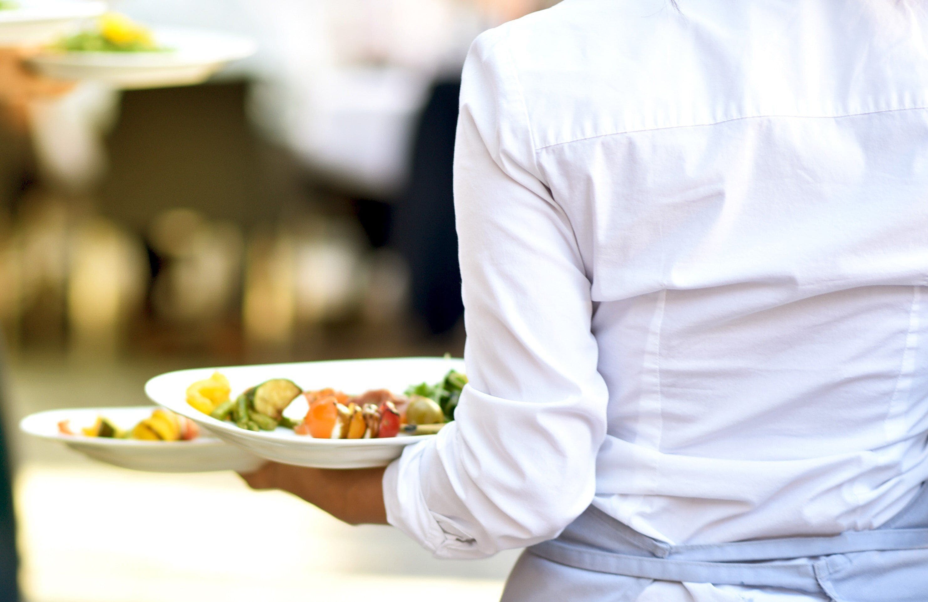 Une personne vêtue d’une chemise blanche tenant des assiettes de nourriture.