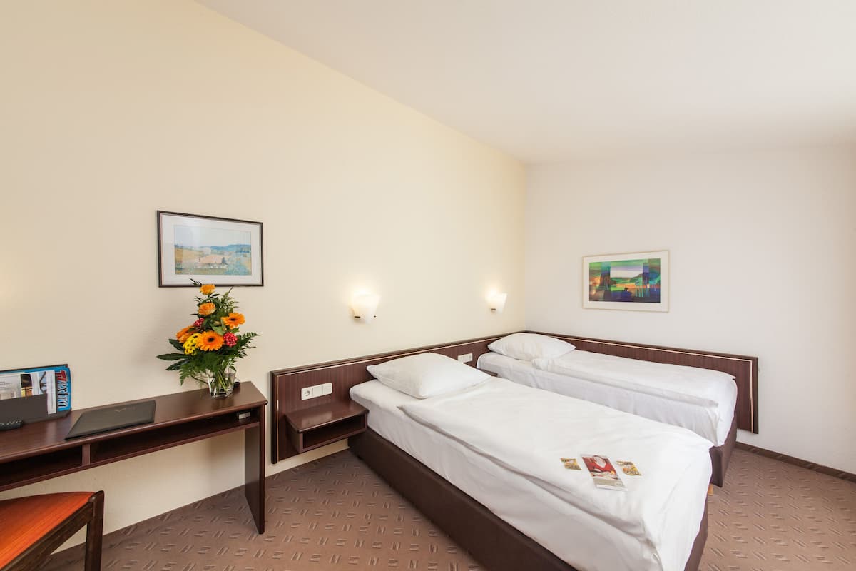 Une chambre d’hôtel avec deux lits simples, un bureau avec un ordinateur portable, un vase de fleurs et deux tableaux encadrés au mur.