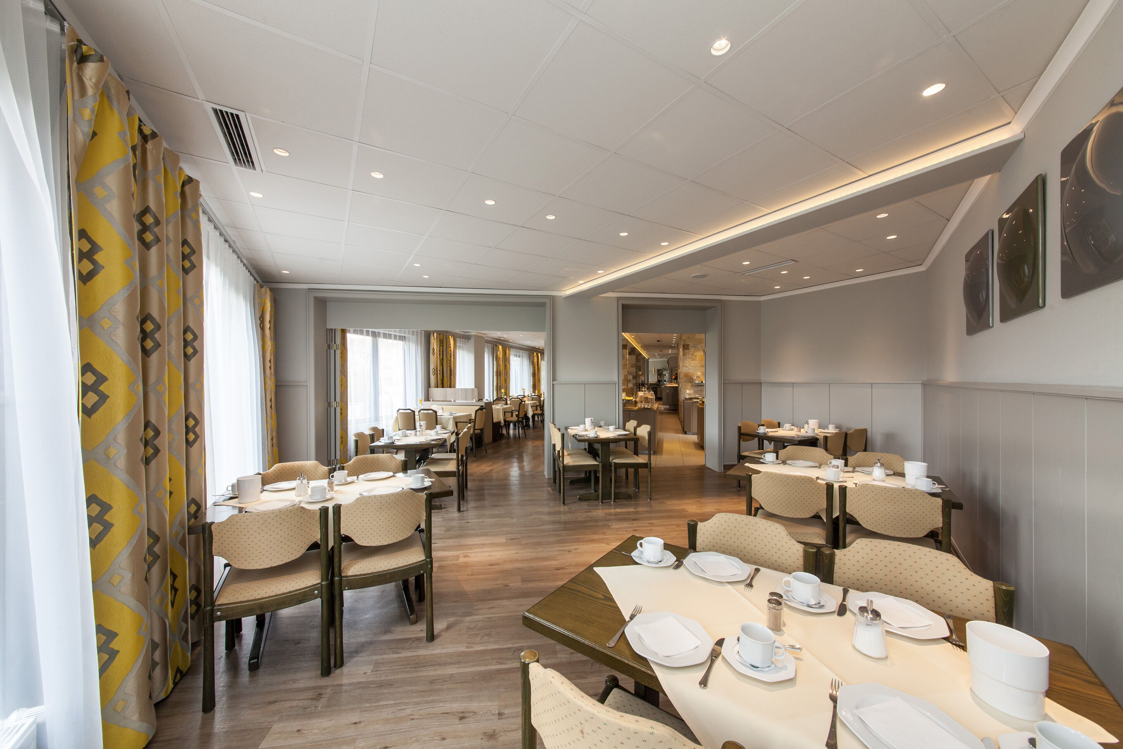A spacious dining area with multiple tables and chairs, a kitchen in the background, and a modern ceiling design.