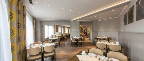 A spacious dining area with multiple tables and chairs, a kitchen in the background, and a modern ceiling design.