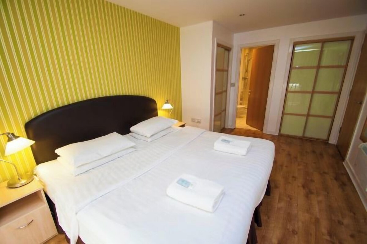 A hotel room with a bed, bedside lamps, a wooden floor, and a door leading to a bathroom.