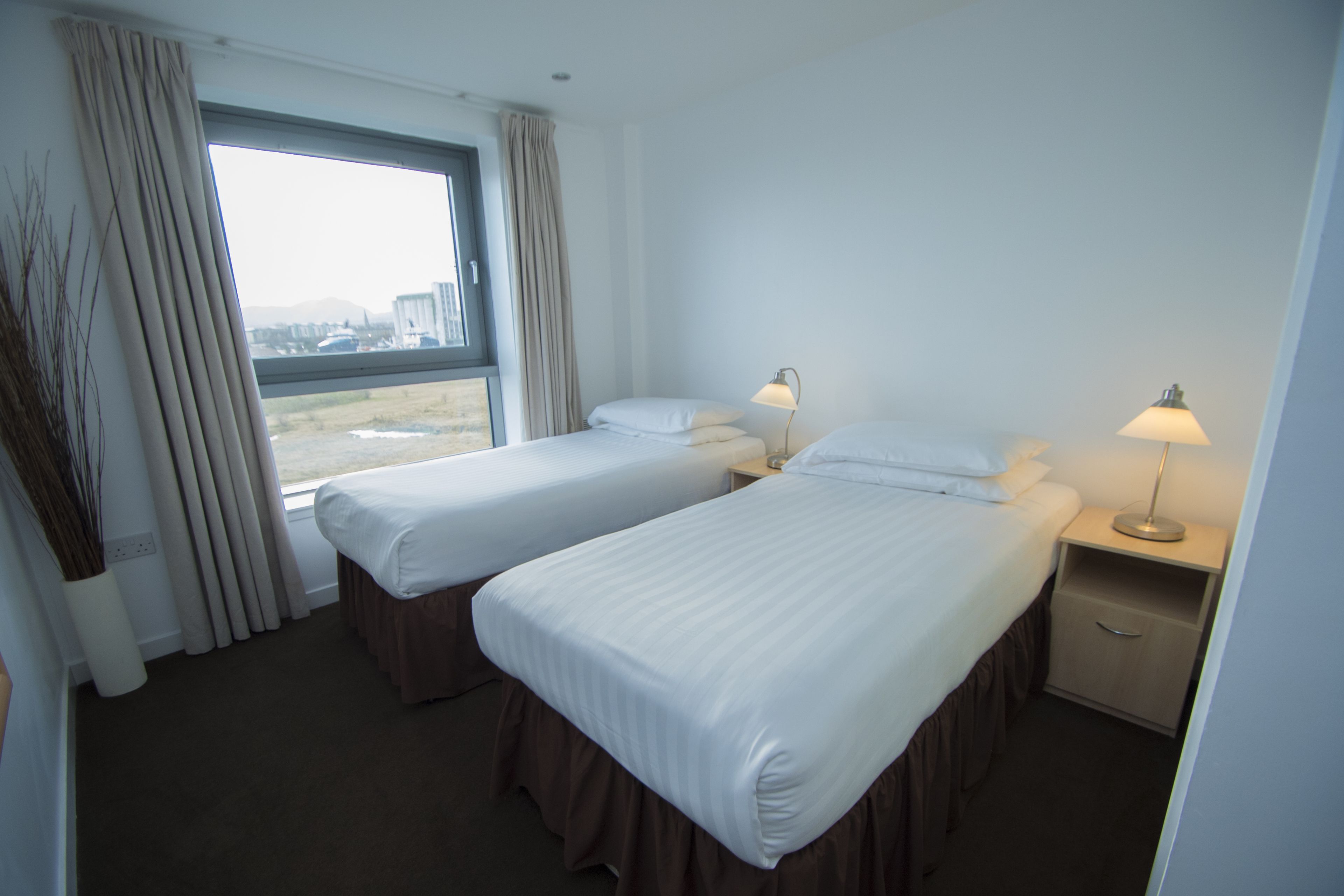 A hotel room with two beds, a window with curtains, bedside lamps, and a view of the outside.