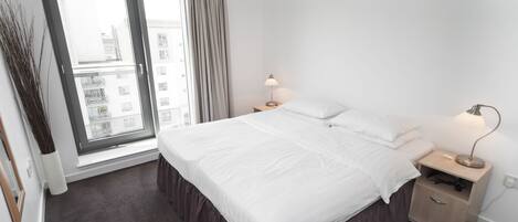 A hotel room with a large bed, a nightstand, a lamp, a window with curtains, and a view of buildings outside.