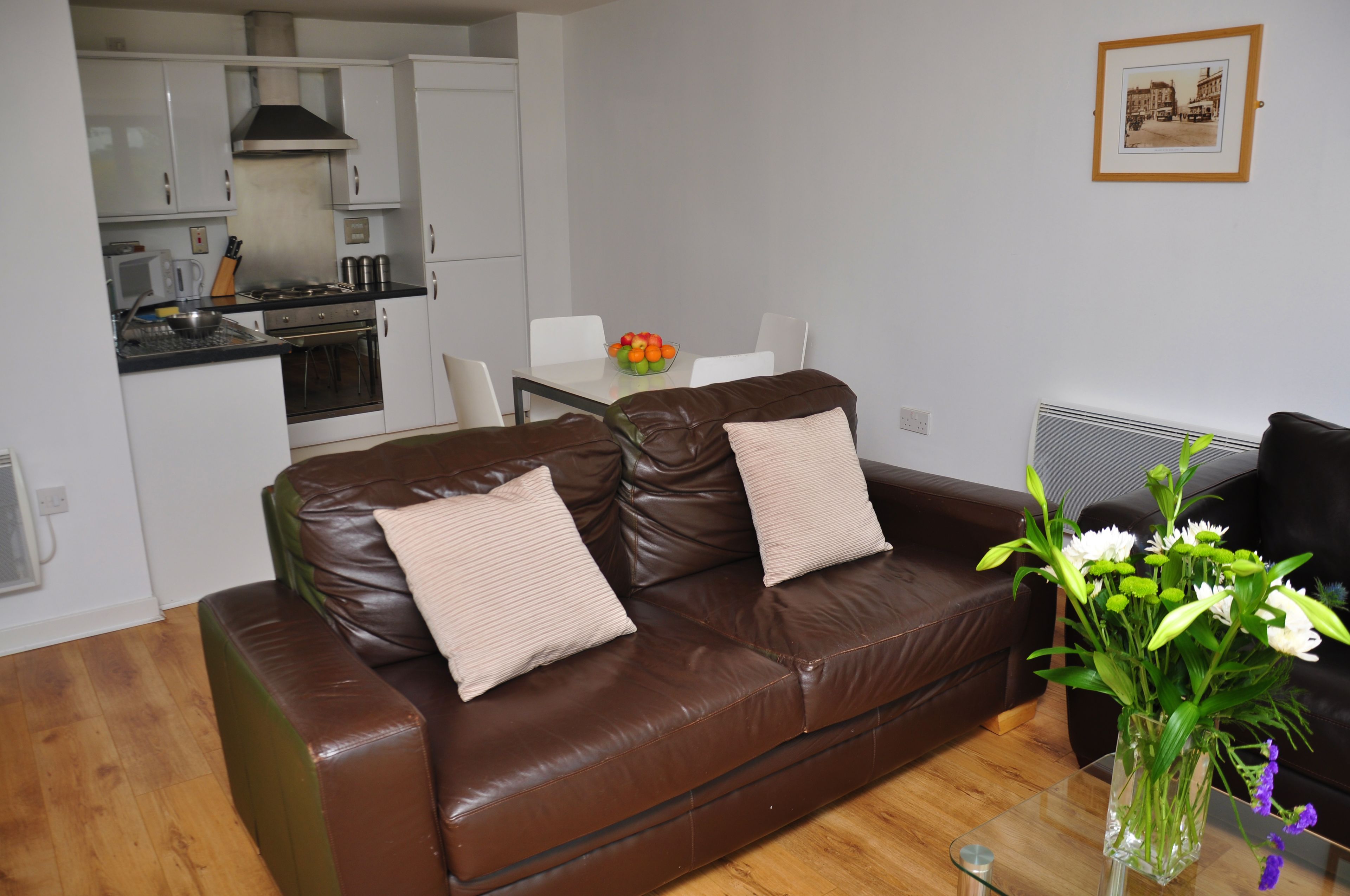 A brown leather sofa with two cushions, a dining table with chairs, a kitchen area, and a vase with flowers on a glass table.