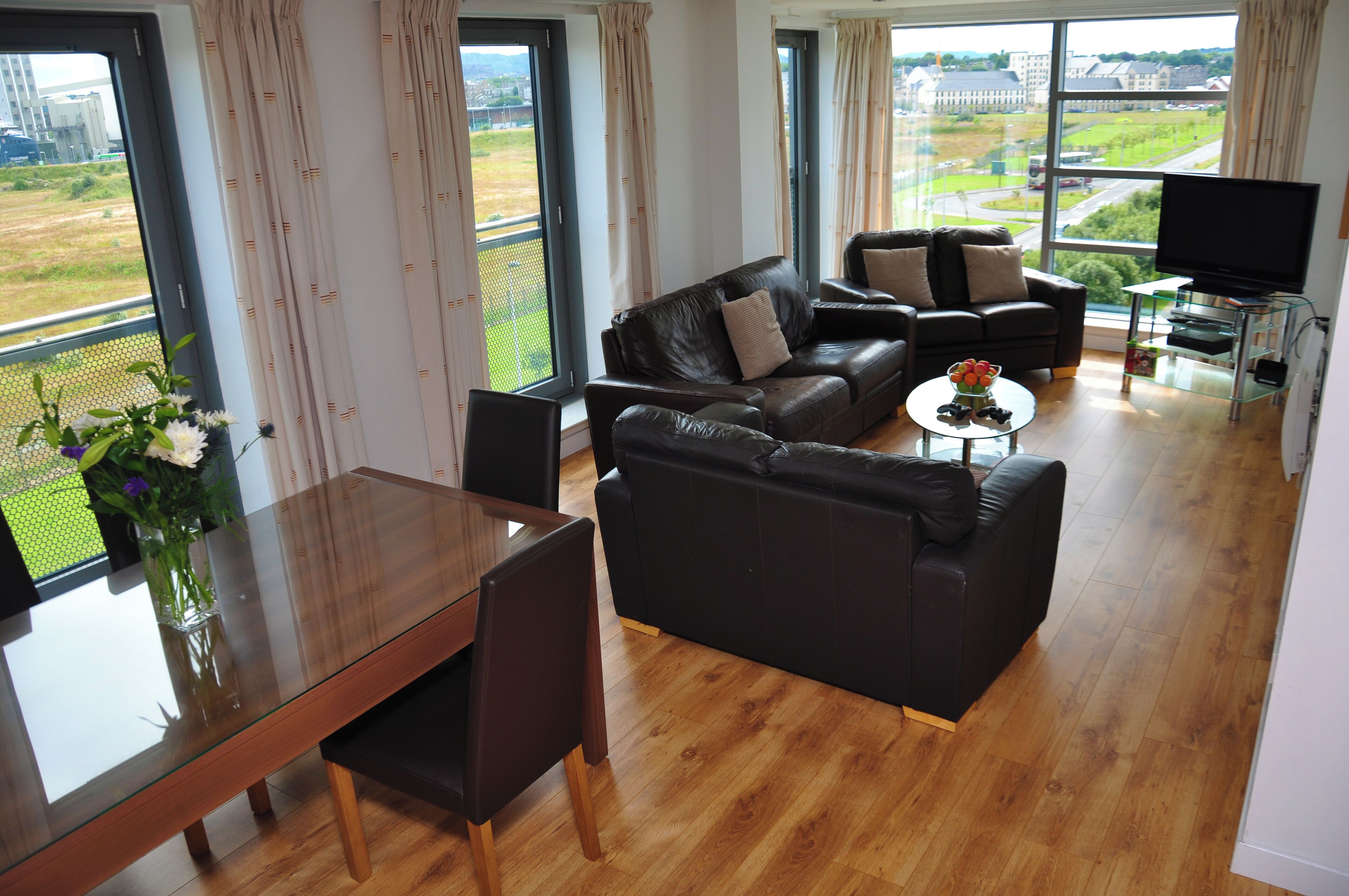 Una sala de estar moderna con una mesa de comedor de vidrio, dos sofás de cuero negro, una mesa de centro de vidrio y un televisor sobre un pedestal de vidrio.