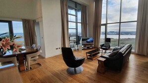 A spacious living room with a large window, a black leather sofa, a coffee table, and a TV. - Ocean Serviced Apartments (Edinburgh)