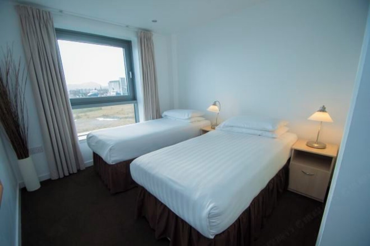 A hotel room with two beds, a window with curtains, a nightstand with a lamp, and a view of the outside.