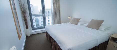 A hotel room with a large bed, a window with curtains, a bedside table, and a view of buildings outside.