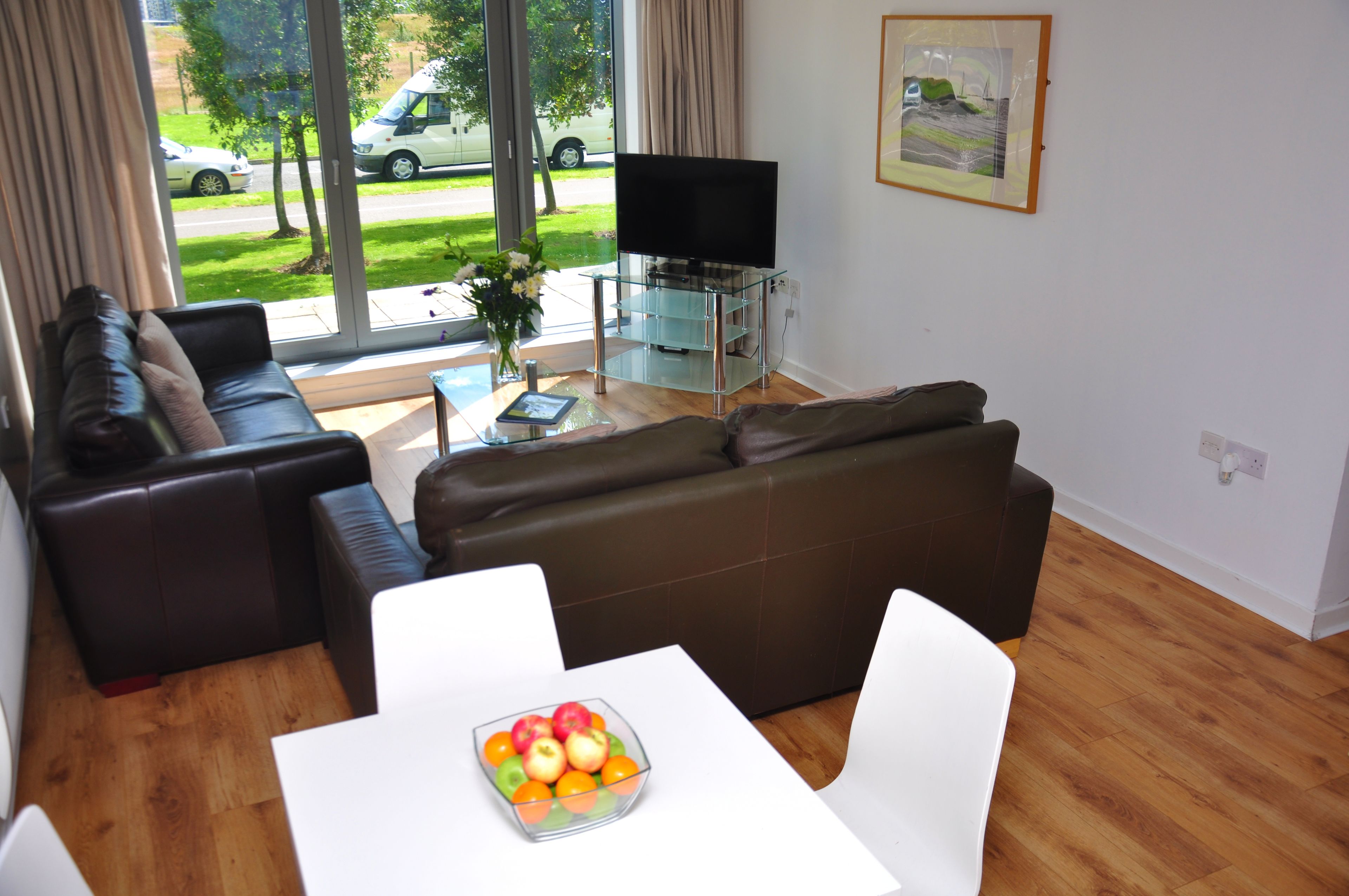 A modern living room with a glass TV stand, a brown leather sofa, a white table with a fruit bowl, and a view of the street through large windows.