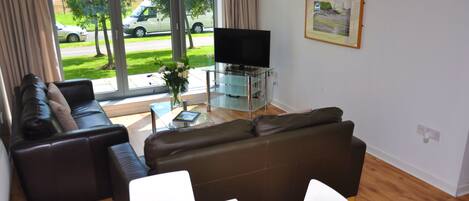 A modern living room with a glass TV stand, a brown leather sofa, a white table with a fruit bowl, and a view of the street through large windows.