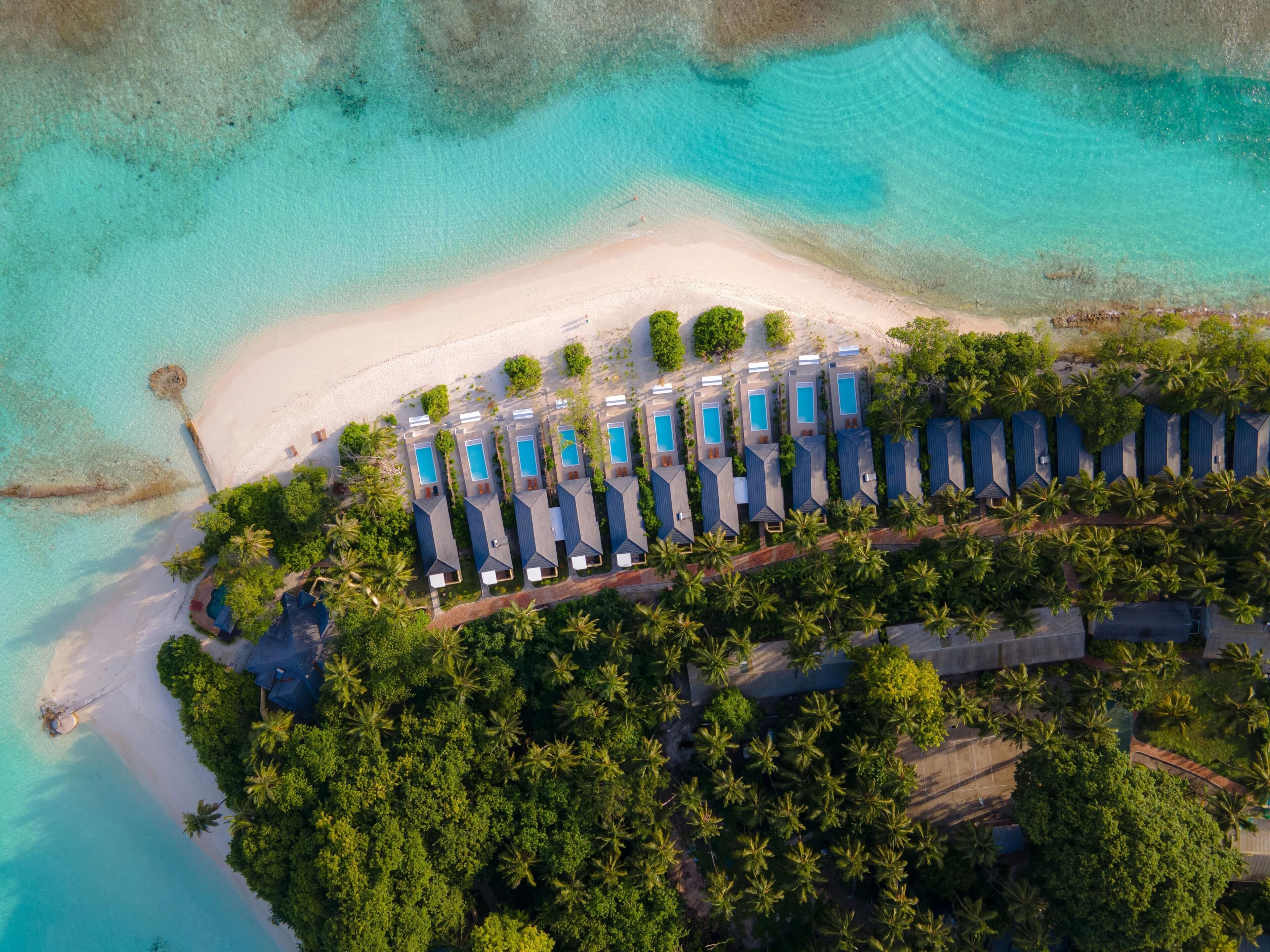 An aerial view of a resort with multiple bungalow-style accommodations, a sandy beach, and clear turquoise waters.