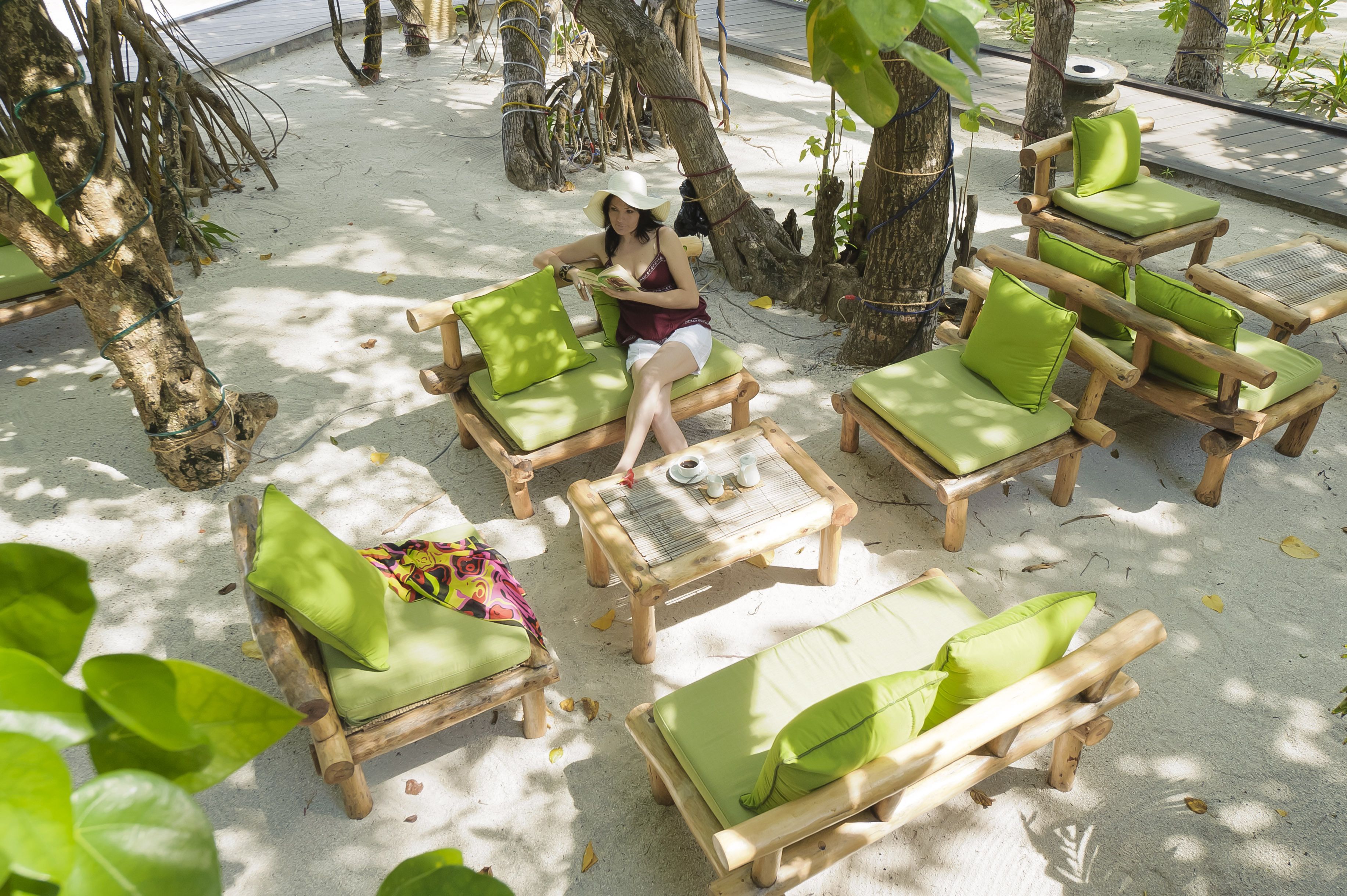 A serene outdoor seating area with wooden lounge chairs and green cushions, surrounded by trees and a sandy ground.