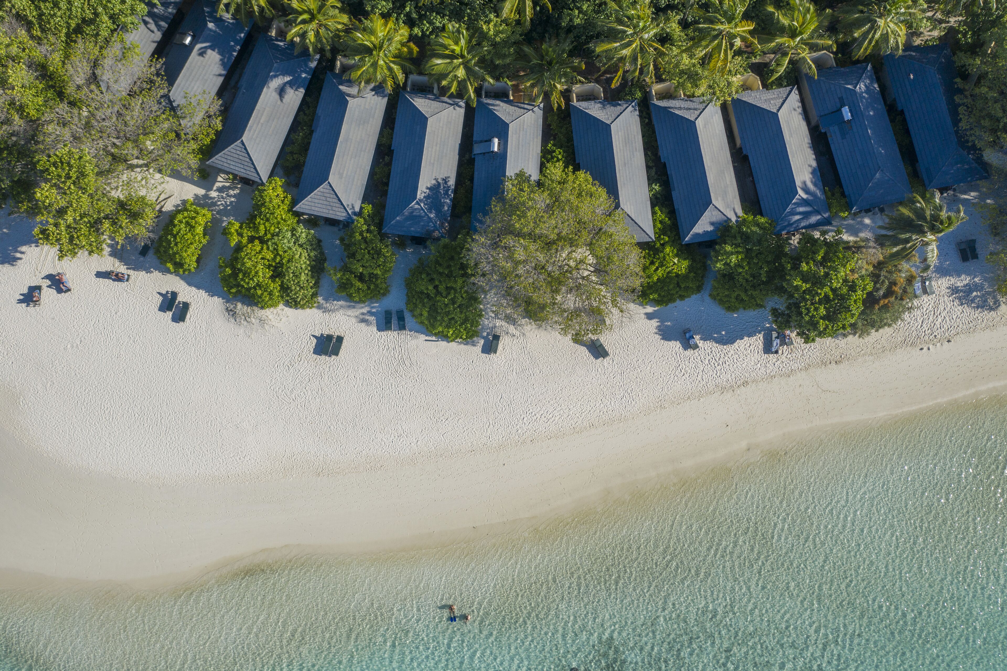 An aerial view of a beachside resort with multiple bungalow-style accommodations, a sandy beach, and palm trees.