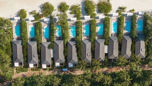 Vista aérea de un resort frente al mar con varias cabañas sobre el agua, cada una con piscina, rodeado de exuberante vegetación y una playa de arena.