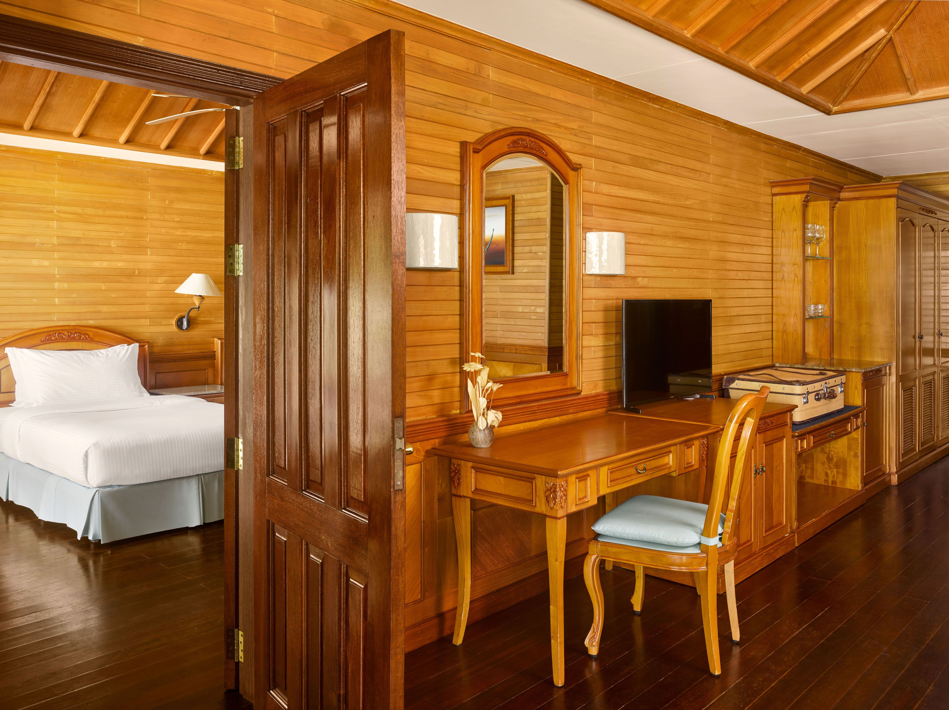 A wooden-finished room with a bed, a desk with a chair, a television, and a fireplace.