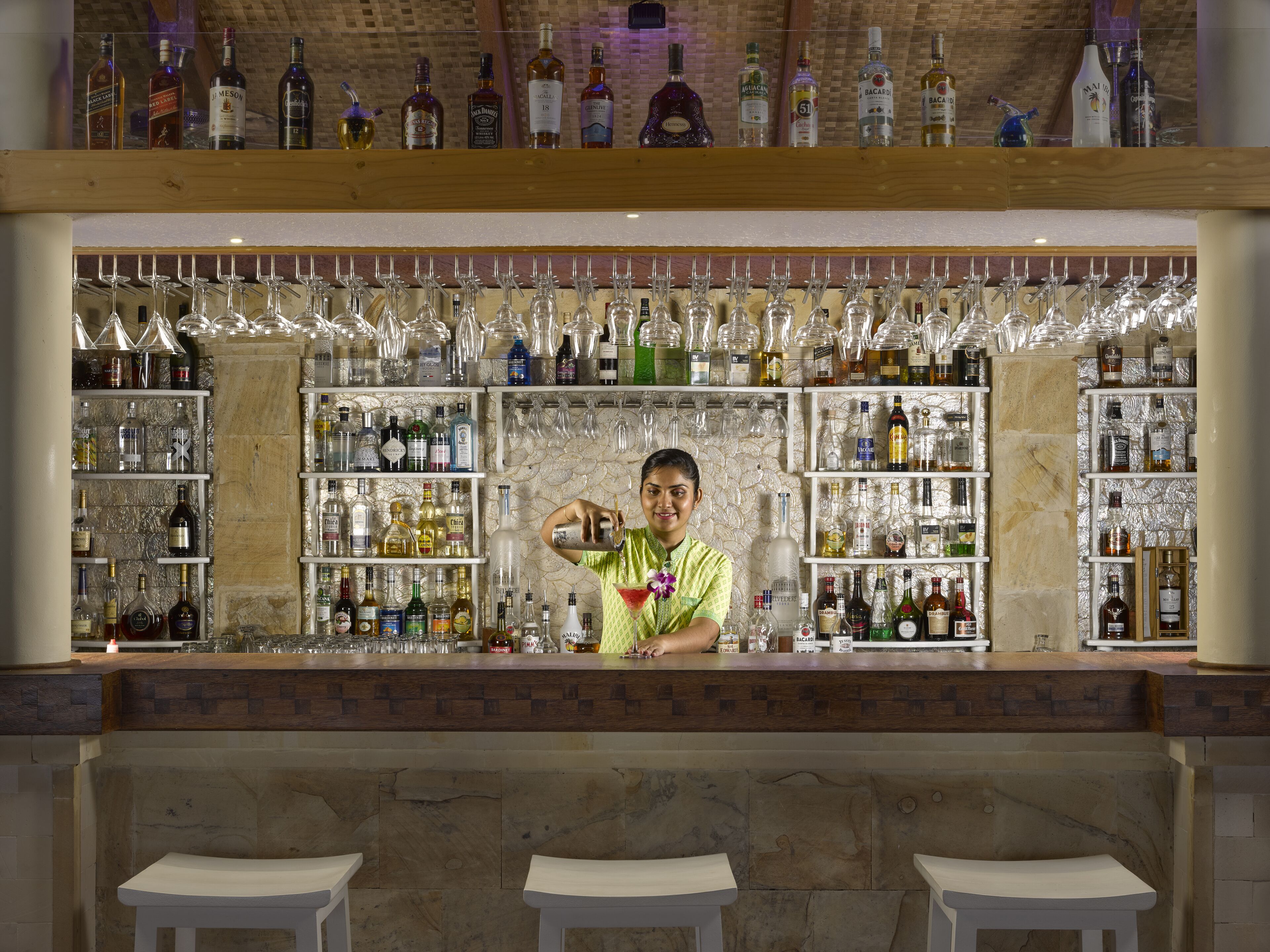 Un bar con un camarero preparando una bebida, botellas de licores en las estanterías y copas de vino colgadas.