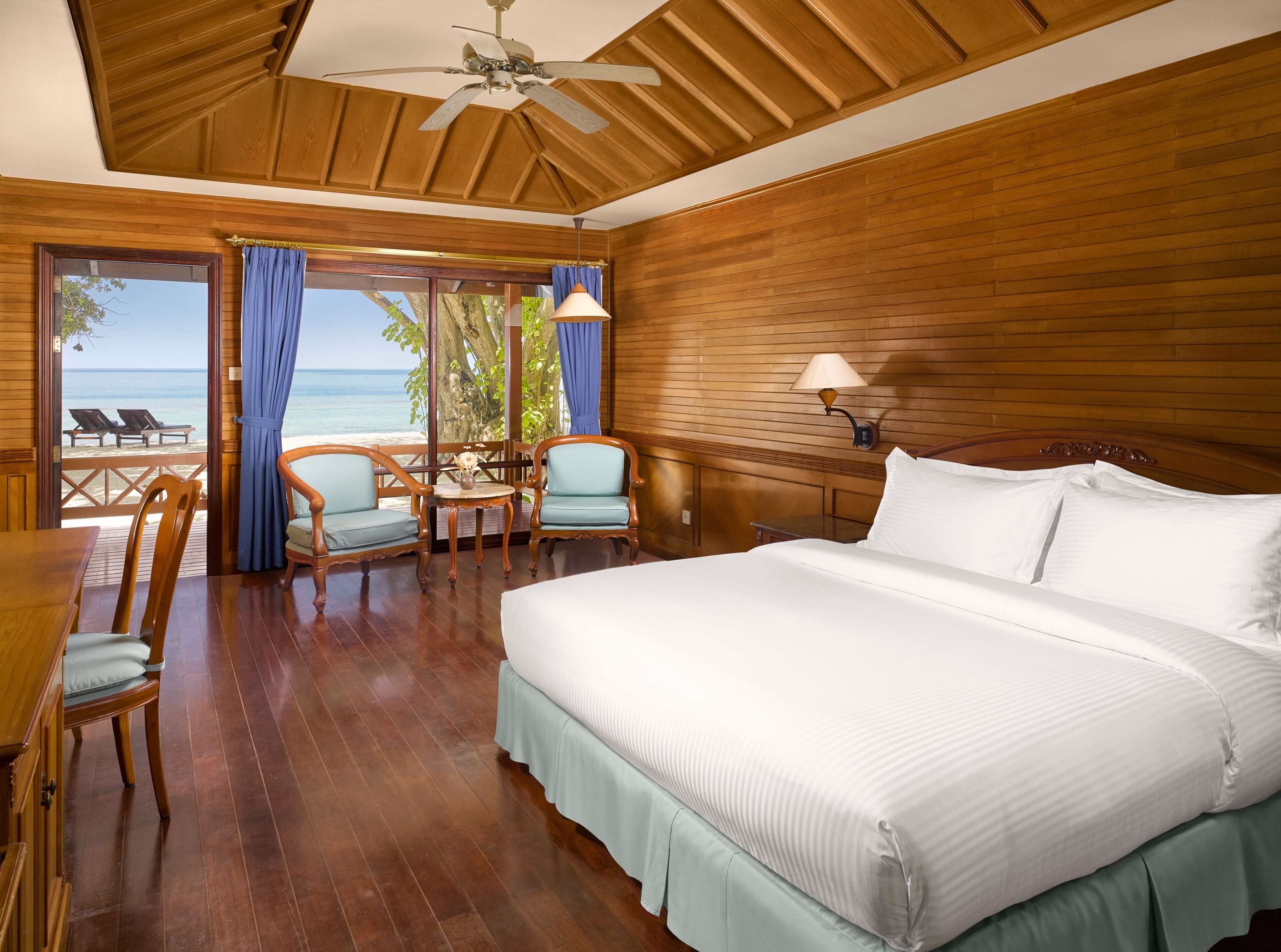A bedroom with a large bed, wooden walls, a ceiling fan, a table with chairs, and a view of the beach.
