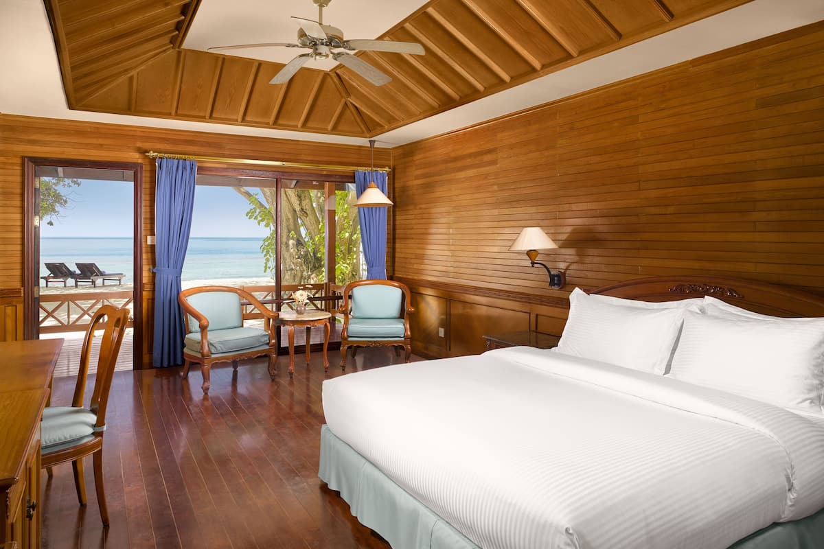 Ein Schlafzimmer mit einem großen Bett, Holzwänden, einem Deckenventilator, einem Tisch mit Stühlen und Blick auf den Strand.