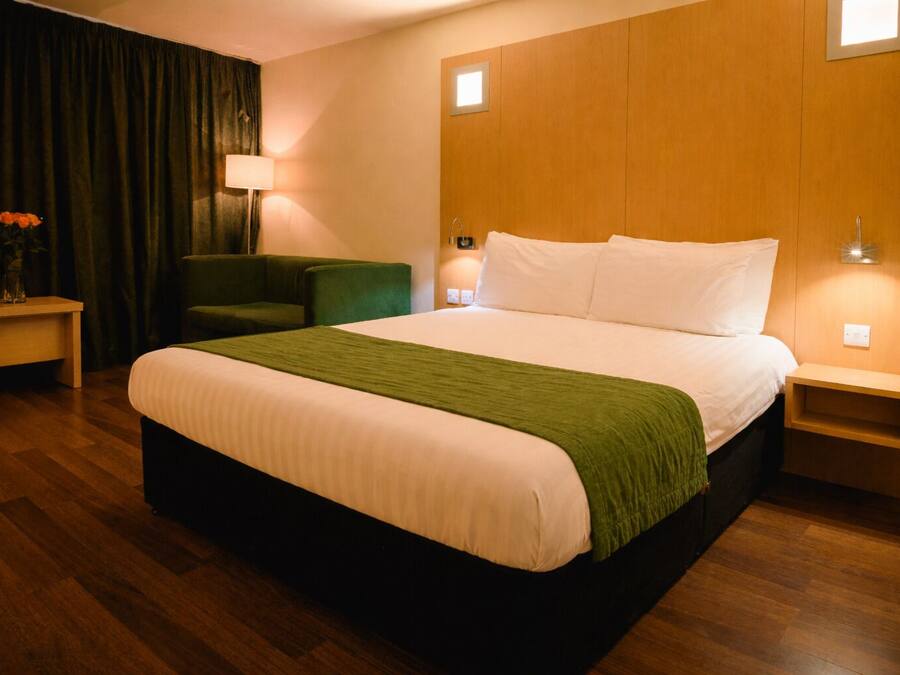 A hotel room with a large bed, a green chair, a nightstand, and a lamp.