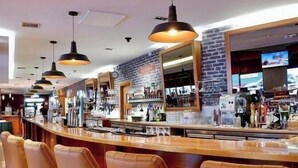 A bar area with leather barstools, pendant lighting, a brick wall, and a counter with various bottles and glasses. - Station House Hotel (Letterkenny)