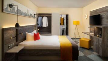 A modern hotel room with a large bed, a bedside table, a TV, a desk, a chair, a lamp, and a wardrobe.