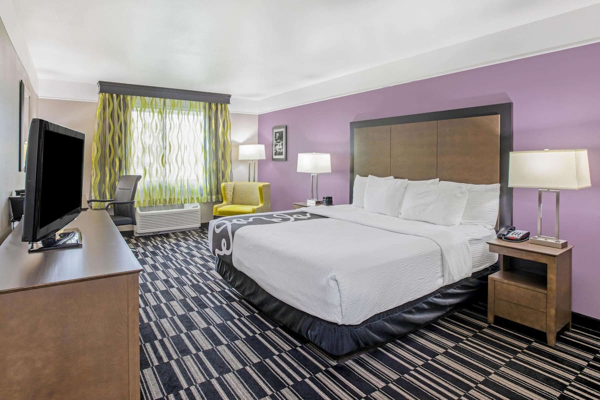 A hotel room with a bed, a desk with a television, a chair, a bedside table with a lamp, and a window with curtains.